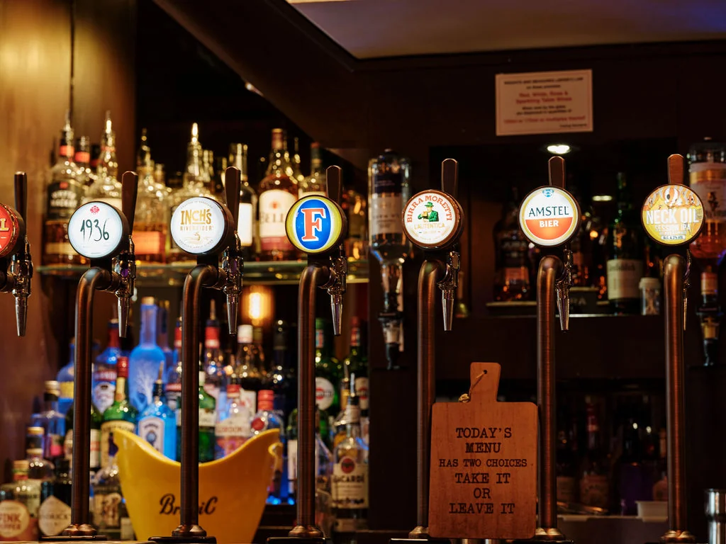 The image shows a well-organized bar with six beer taps, each with a unique label. The taps are arranged in a row, with the largest tap on the left and the smallest on the right. The background features a shelf of various liquor bottles, and a wooden sign with the text "Brewery" is visible. The scene is illuminated by warm, ambient light, creating a cozy atmosphere.