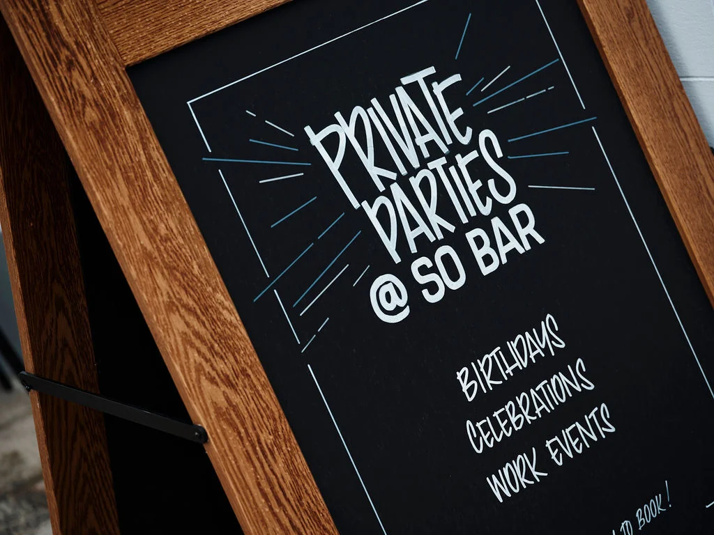 A black chalkboard sign with white text and a black frame is displayed on a wooden stand against a white wall. The sign advertises a "Piano & Piano Bar" with "Piano Bar 10am - 10pm" and "Piano Bar 11am - 11pm". The bar offers "Piano Bar Music", "Piano Bar Music", and "Piano Bar Music". The sign is positioned on the left side of the frame.