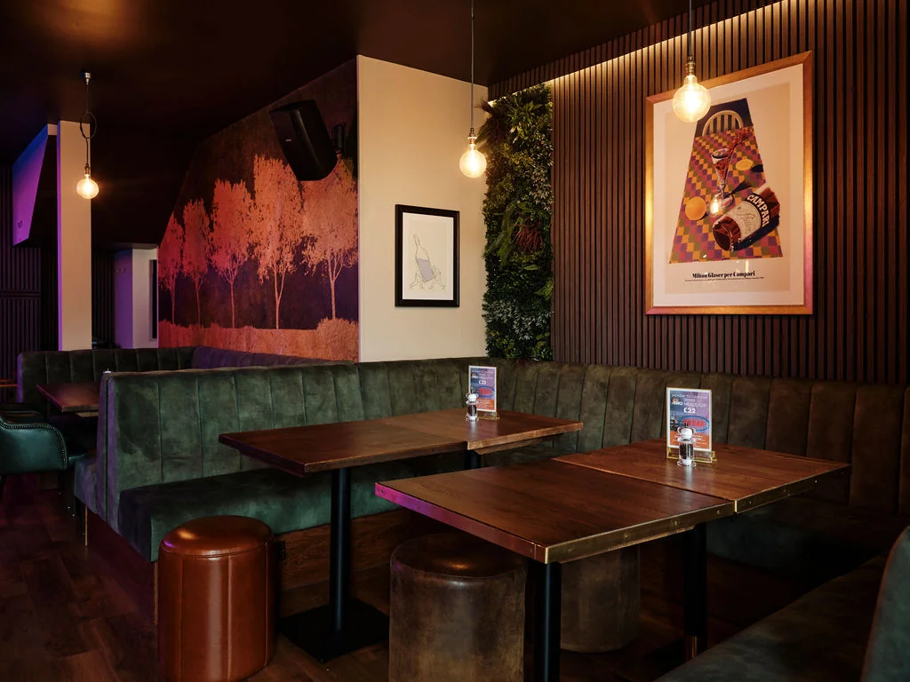 A modern restaurant interior features a wooden floor, green booths, and a wooden bench. The walls are adorned with colorful posters and a large poster with a tree design. The ceiling has hanging lights and a black speaker. The atmosphere is cozy and inviting, with a warm ambiance.