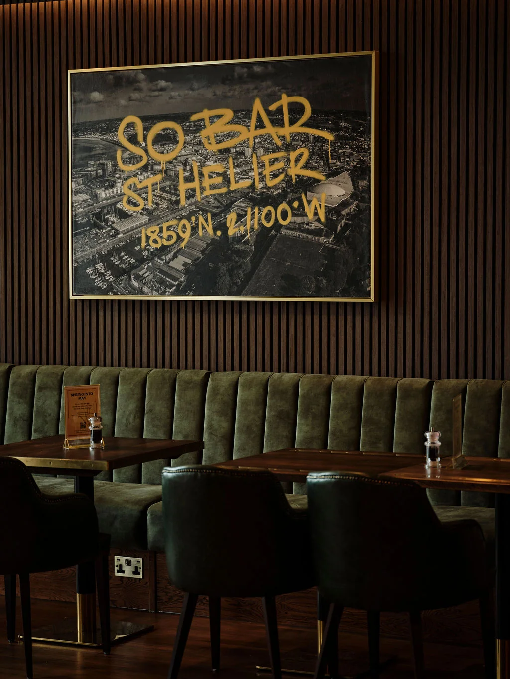 A modern restaurant features a large black and white poster with "SOFT BEER" in yellow, advertising a special offer for "SOFT BEER 20% OFF". The poster is hung on a wooden wall, with a green booth and wooden bar in front of it. The floor is dark wood, and the overall atmosphere is warm and inviting.