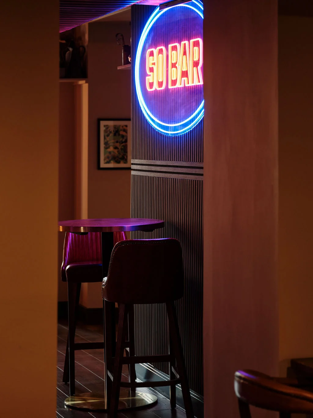 A dimly lit, cozy bar scene features a neon "SAND" sign on a white wall, with two wooden stools and a wooden bar in the foreground. The floor is dark wood, and a painting hangs on the wall. The perspective is from the entrance, inviting patrons to join.