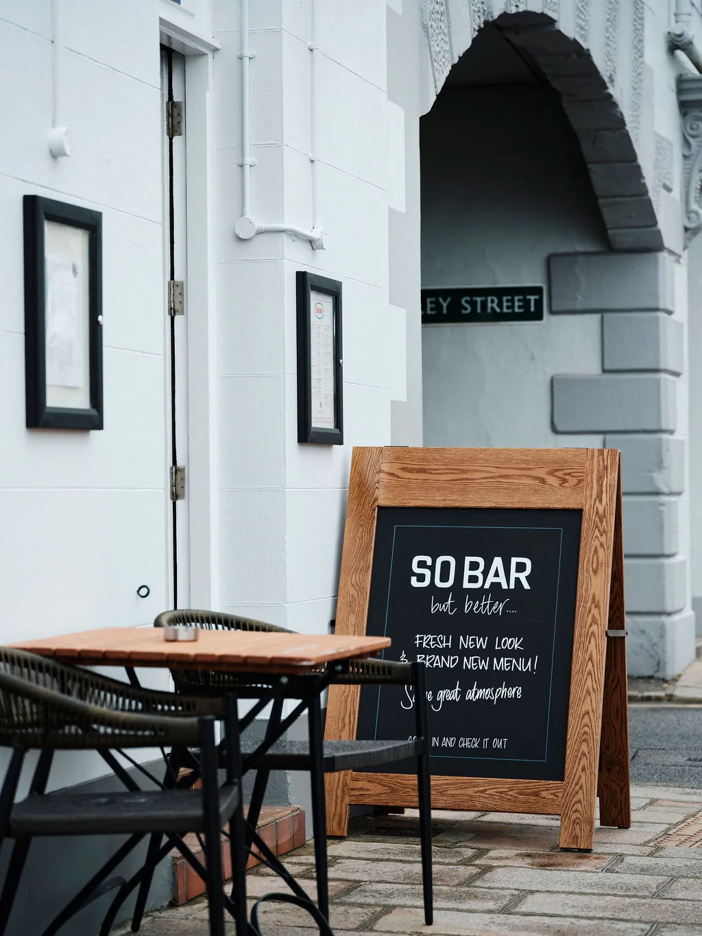 A charming outdoor cafe with a white building as the backdrop features a black chalkboard sign advertising "Soda" and a table with chairs and a glass. The scene is set on a cobblestone sidewalk, with a door in the background.