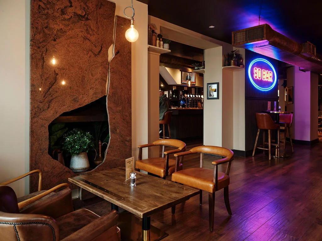 A cozy bar or restaurant features a rustic wooden wall with a large hole, a wooden table with a vase of flowers, and a wooden chair. The bar has a neon sign and a potted plant. The floor is dark wood, and the ceiling has hanging lights. The atmosphere is warm and inviting, with a welcoming ambiance.