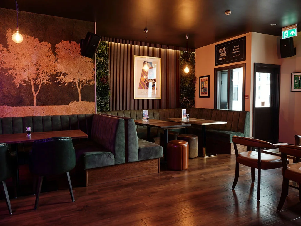 A cozy, rustic restaurant with dark wood floors and green velvet booths is illuminated by hanging lights. A large mural of a tree and a menu sign are on the walls, while a wooden table with a candle and a brown trash can are in the center. A door with a window and a green sign are on the right side, and a hanging light fixture is on the left. The atmosphere is warm and inviting, perfect for a cozy evening.