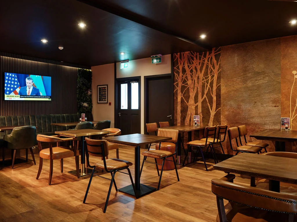 A modern, cozy bar with dark wood floors and a wooden wall features a large flat-screen TV displaying a woman in a blue shirt. The bar has six tables, two green couches, and a wooden bar. A wooden tree mural adds a touch of nature. The bar is well-lit, creating a warm and inviting atmosphere.