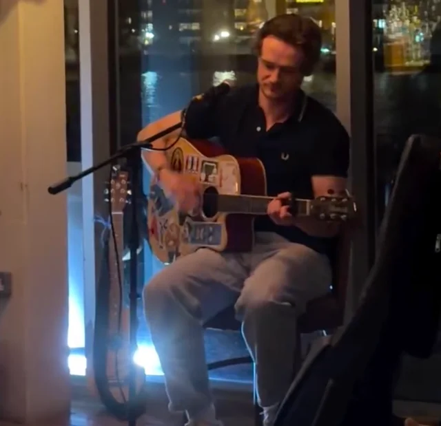 A man in a black polo shirt and blue jeans is playing a guitar in a cozy room with a window overlooking a cityscape. He is seated on a chair with a guitar on his lap, and a microphone is nearby. The room has a warm, inviting atmosphere with a wooden floor and a wooden chair. The man's focused expression and the guitar's sound create a serene and intimate atmosphere.
