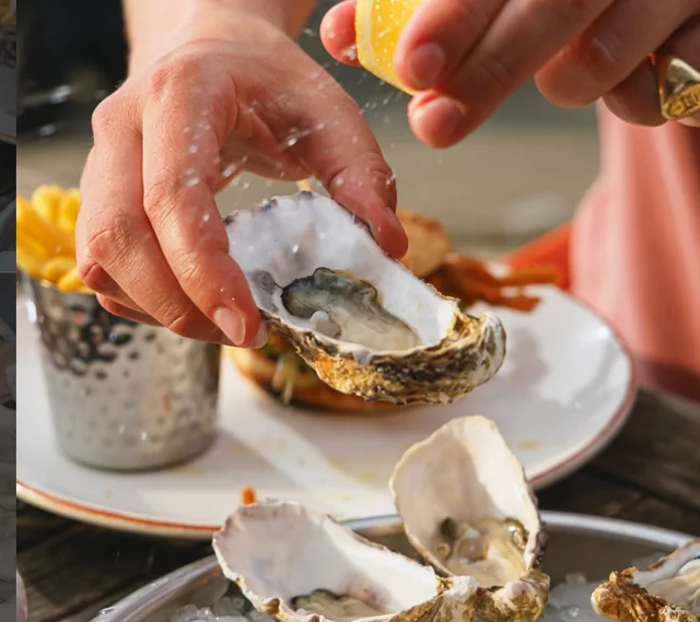 A person in a pink shirt is pouring salt over an open oyster on a white plate, with a silver bucket of fries nearby. The scene takes place on a wooden table, with a blurred background suggesting an outdoor setting.