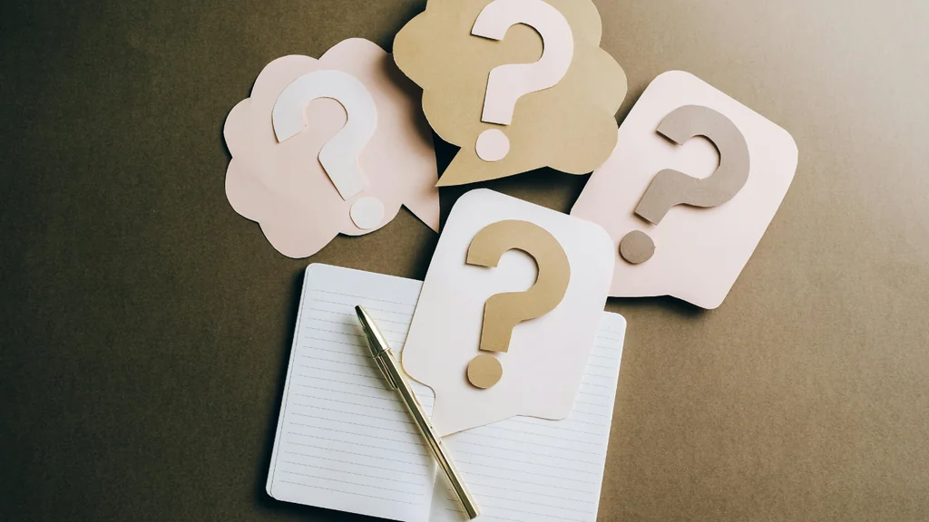 A notebook with a gold pen lies on a dark brown surface, surrounded by six paper bubbles with question marks, two of which are pink and two are beige. The scene suggests a brainstorming session or a problem-solving process.