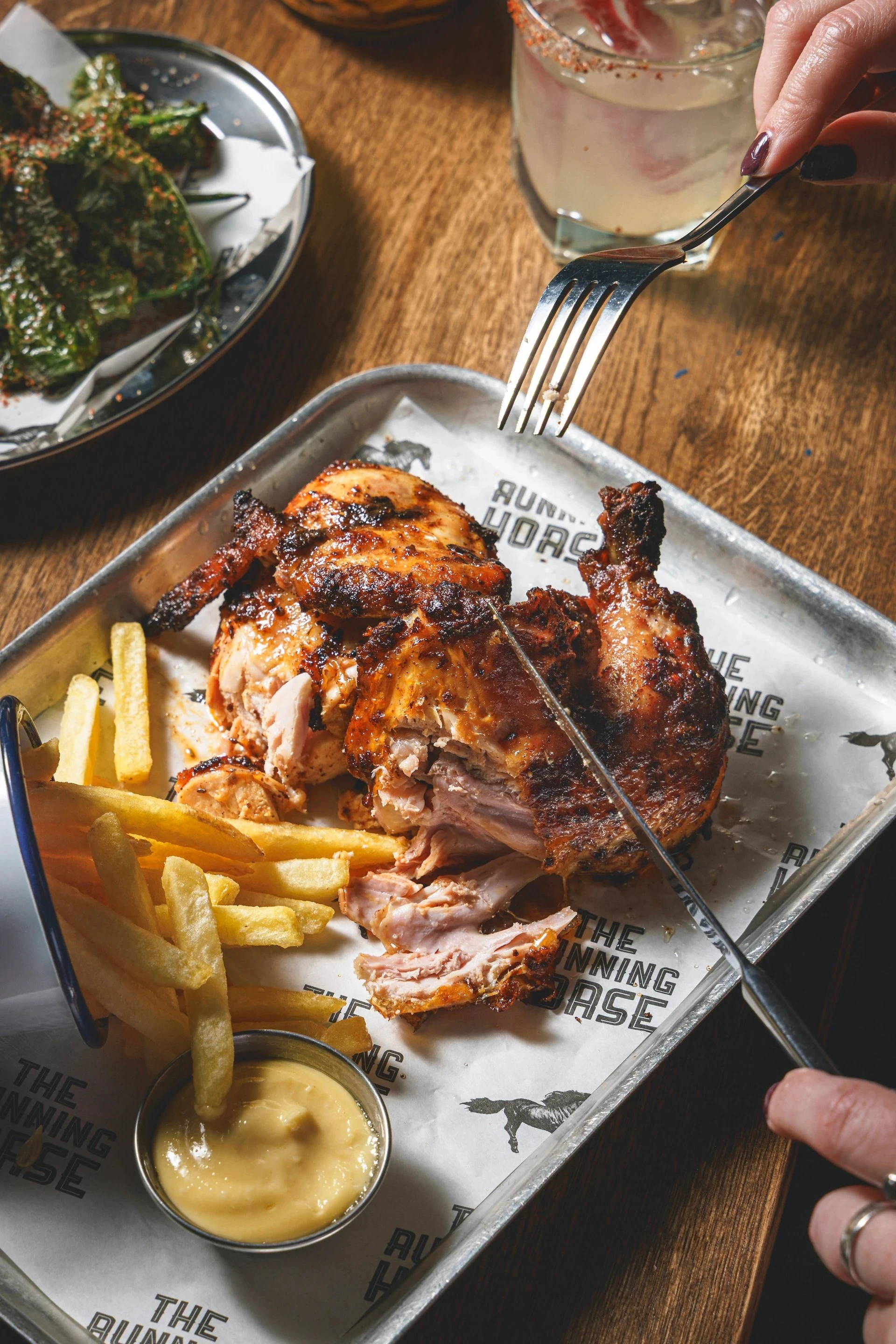 A dining scene features a large metal tray with a grilled chicken, french fries, and a side of sauce. A fork is in the chicken's mouth, and a hand holds a knife. A glass of wine is nearby. The table has a wooden surface and a salad in the background. The image portrays a meal in progress, with the chicken as the main dish.
