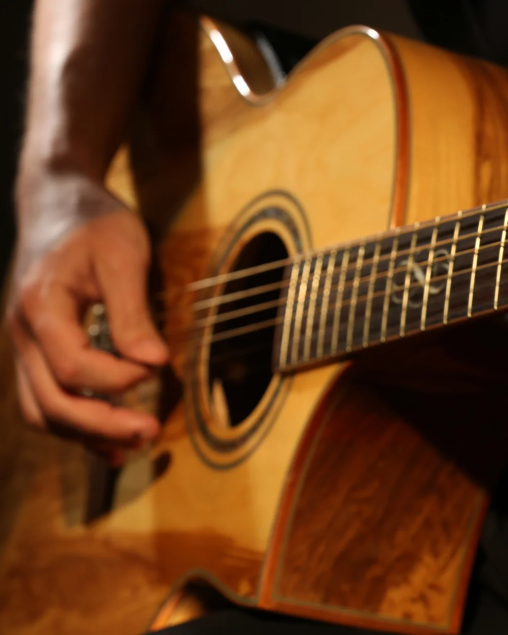 A close-up image shows a hand playing an acoustic guitar, with the guitar's light-colored body and dark strings contrasting against a black background. The hand is positioned on the lower fret, and the guitar's head rests on the neck. The image is taken from a slightly elevated angle, emphasizing the guitar's size and the hand's position. The focus is on the guitar and hand, with no other objects or text present.