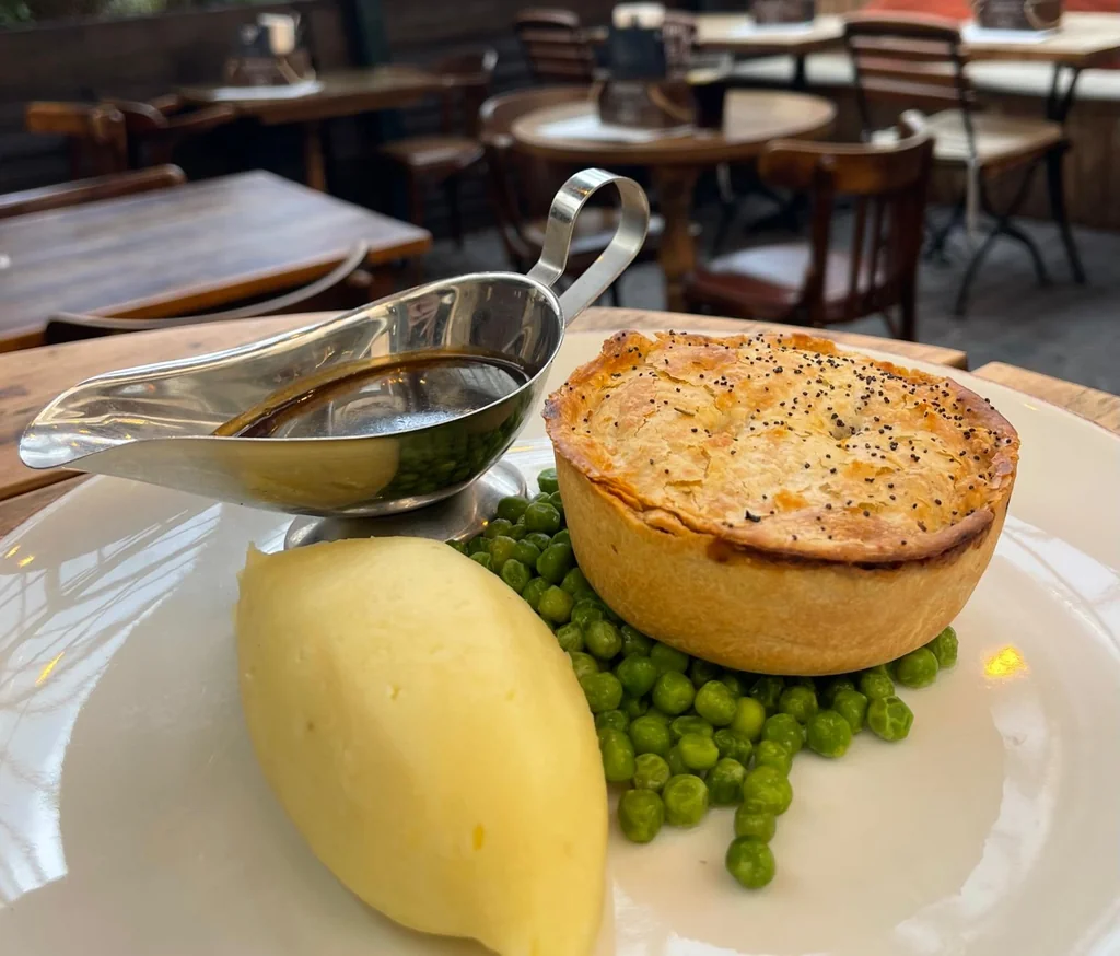 A wooden table in a restaurant features a golden-brown pie with a sprinkle of herbs, a creamy sauce, a roasted potato, and green peas. A silver serving bowl and a metal ladle are nearby, ready for use. The background is blurred, emphasizing the meal's presentation.