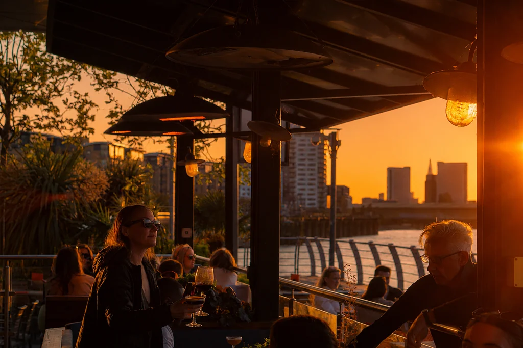 At a rooftop restaurant overlooking a river, people enjoy a sunset view. The restaurant has a black metal roof and a metal railing. The city skyline is visible in the background, and the warm orange sky casts a golden glow. The scene is serene and inviting, with people engaged in conversation and enjoying the view.